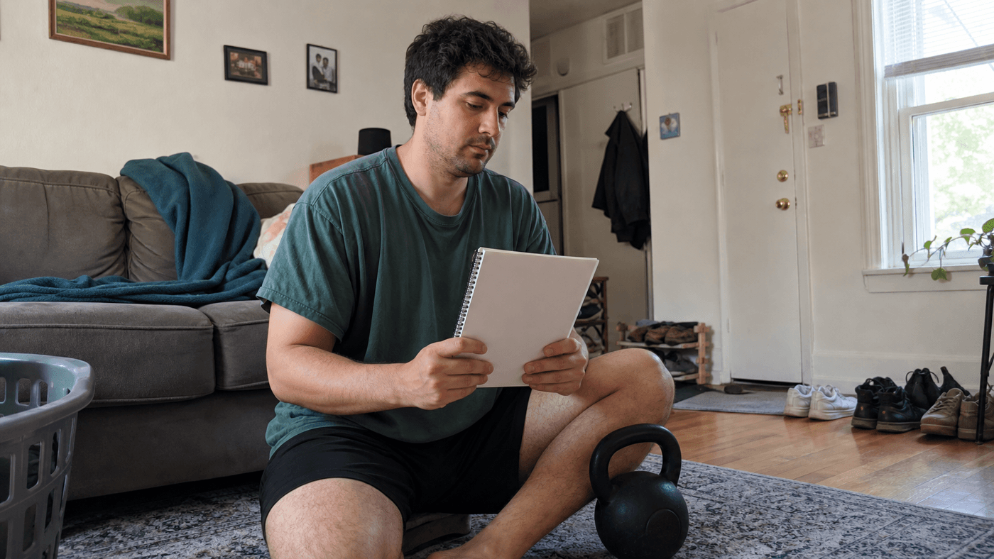 Adult reviewing a blank notebook beside a kettlebell in a lived-in apartment living room