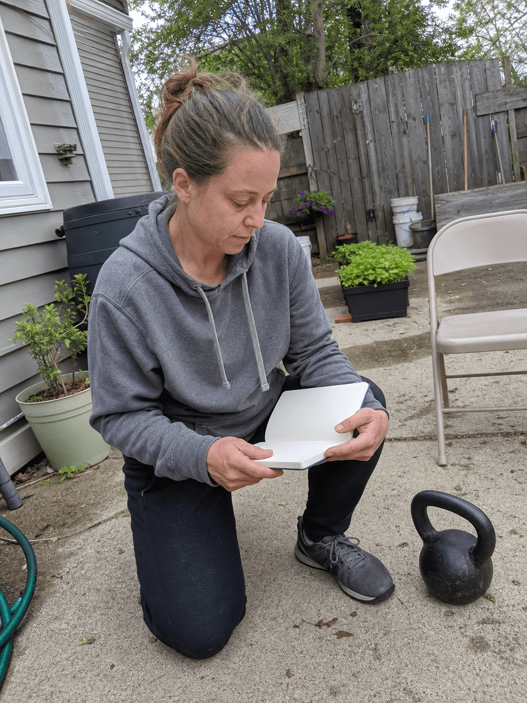 Adult reviewing a blank notebook beside a kettlebell on a lived-in backyard patio