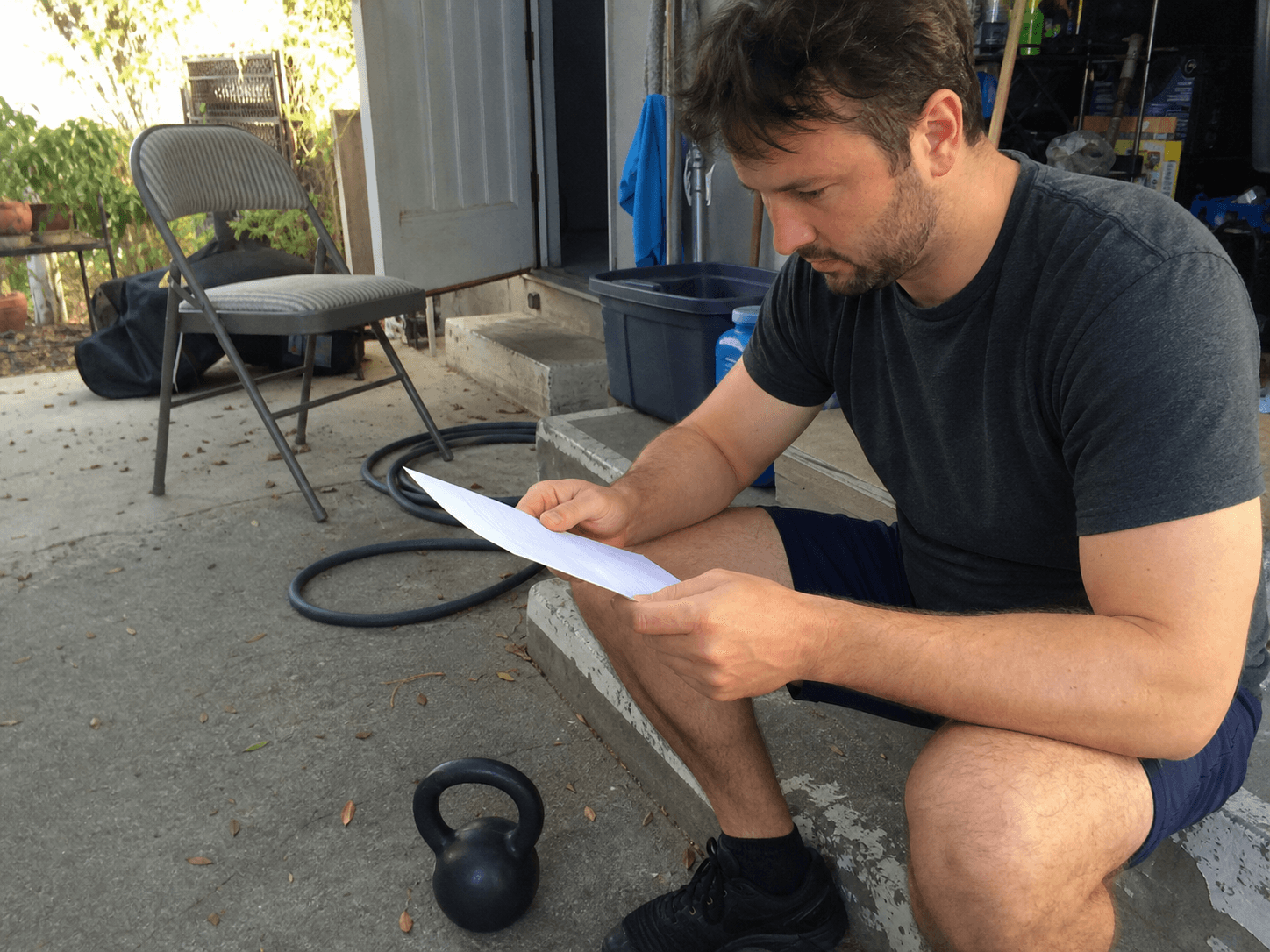 Adult reviewing a blank training check-in beside a kettlebell at a lived-in carport doorway