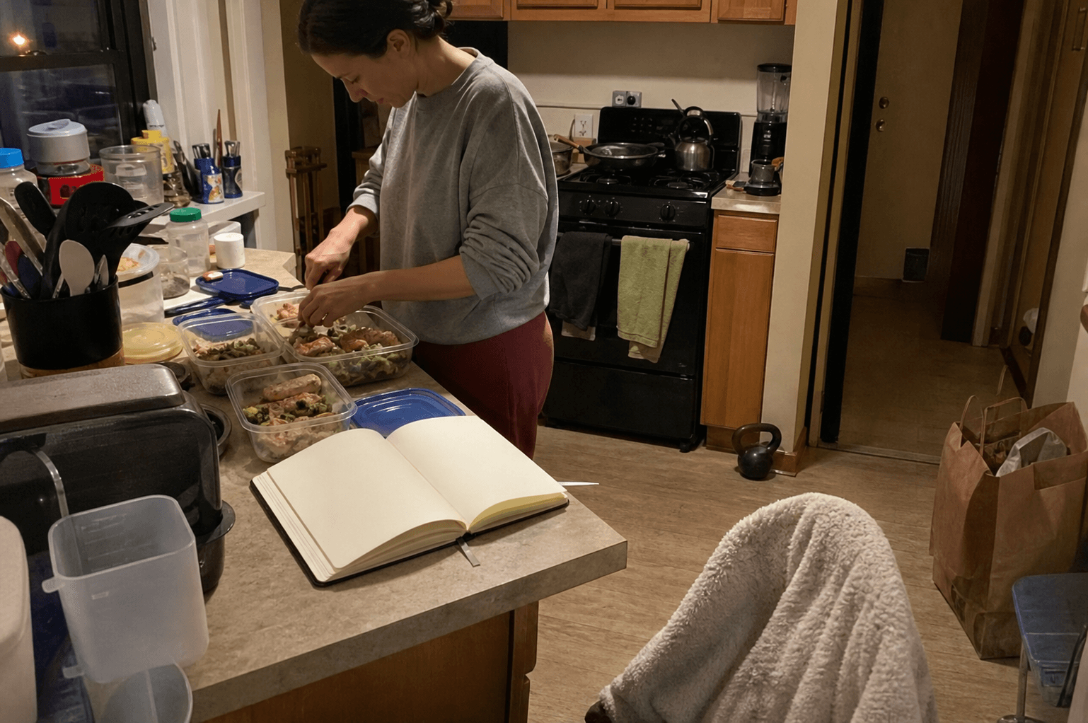 Adult preparing simple meals beside a blank notebook in a lived-in apartment kitchen